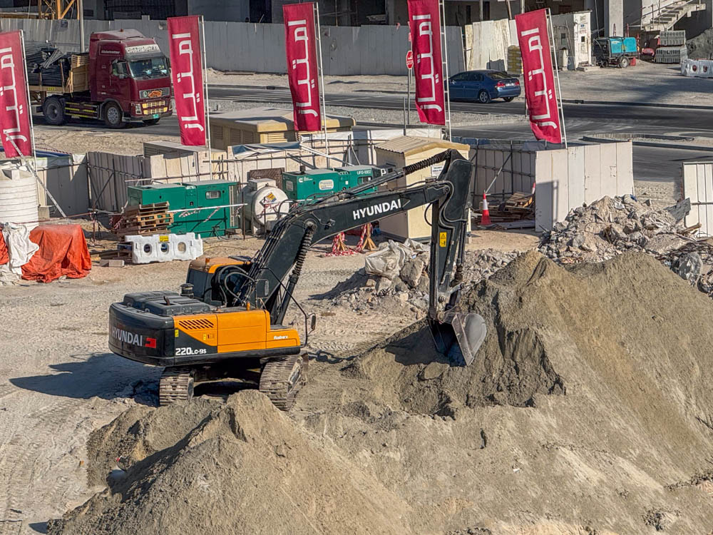 Cotier House 2 By Imtiaz A construction site featuring a Hyundai excavator digging into a pile of dirt, with trucks and banners in the background.